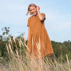 May include: A woman in a brown linen dress smiles while standing in a field of tall grass.