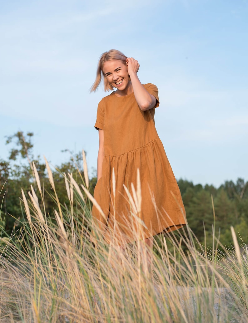 May include: A woman in a brown linen dress smiles while standing in a field of tall grass.