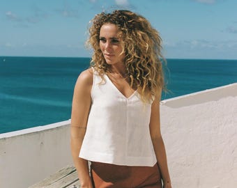 May include: A woman wearing a white sleeveless linen top with a v-neck and a brown skirt. She is standing in front of a white wall and a wooden railing with the sea in the background.