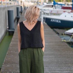 May include: A woman wearing a black sleeveless linen top and olive green linen trousers. She is standing on a wooden jetty with boats in the background.
