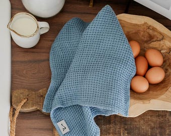 Può includere: Un asciugamano da cucina in tessuto a nido d'ape blu con un piccolo cartellino che dice "~" su un tagliere di legno. L'asciugamano è piegato e drappeggiato sul tagliere. Ci sono uova marroni in una ciotola di legno dietro l'asciugamano.