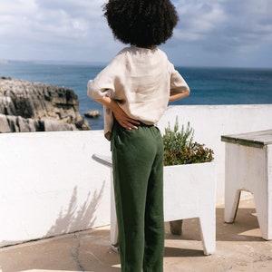 May include: A person wearing a beige linen top and dark green linen pants stands on a white patio overlooking the ocean. The outfit is paired with brown sandals. The scene is set against a backdrop of a blue sky and rocky coastline.