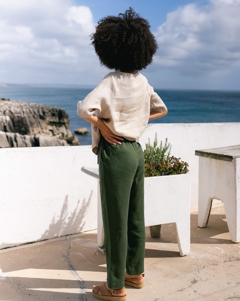 May include: A person wearing a beige linen top and dark green linen pants stands on a white patio overlooking the ocean. The outfit is paired with brown sandals. The scene is set against a backdrop of a blue sky and rocky coastline.