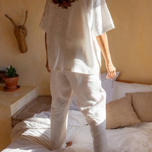 以下が含まれることがあります： 白いリネン素材のパジャマを着た人が、白いベッドの上に立っています。ベッドには白いシーツと枕が敷かれています。