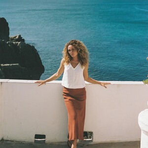 May include: A woman wearing a white V-neck top and a rust-colored maxi skirt stands on a white wall overlooking the ocean. The woman has curly hair and is wearing sandals. The background features a clear blue sky and ocean.