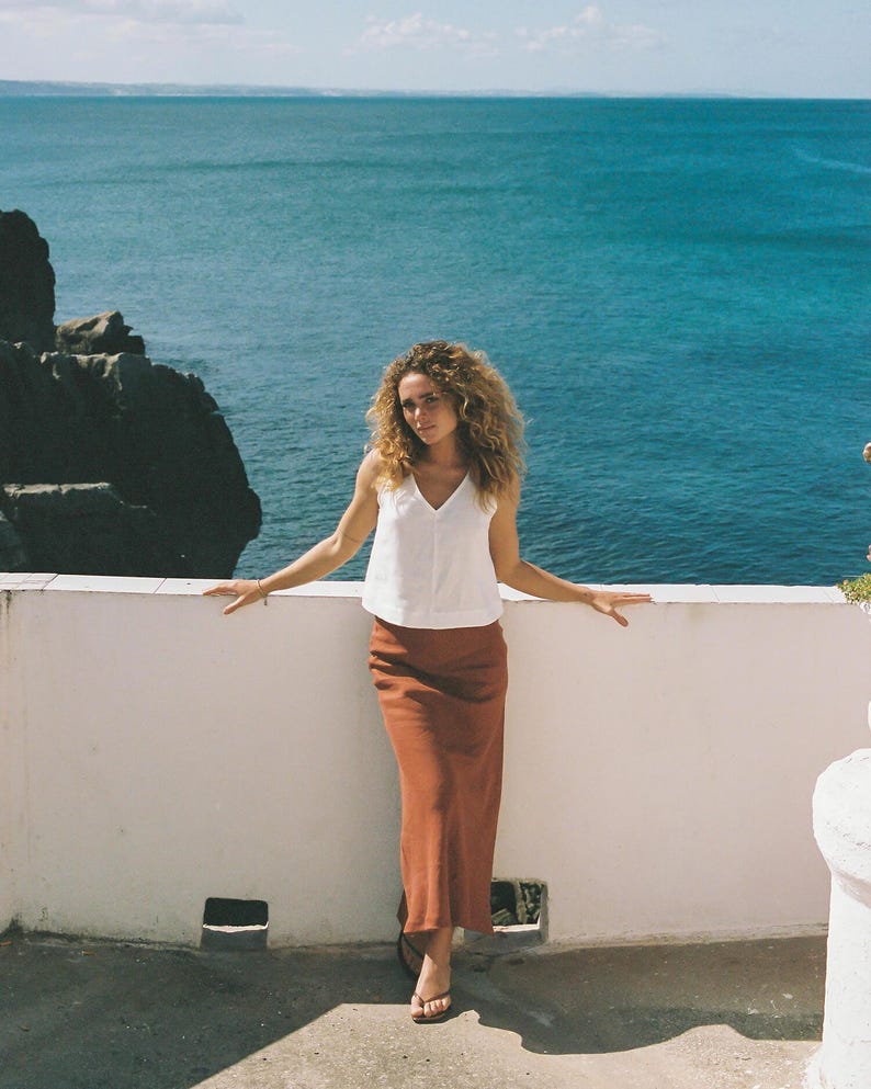 May include: A woman wearing a white V-neck top and a rust-colored maxi skirt stands on a white wall overlooking the ocean. The woman has curly hair and is wearing sandals. The background features a clear blue sky and ocean.