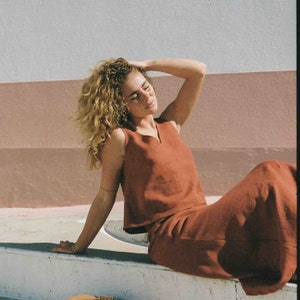 May include: A woman wearing a rust-colored sleeveless linen top and matching skirt. She is sitting on a white surface with her eyes closed and her hand on her head.
