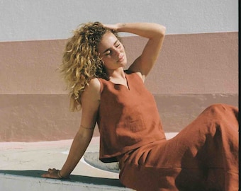 May include: A woman wearing a rust-coloured sleeveless linen top and matching skirt. She is sitting on a white surface with her eyes closed and her hand on her head.