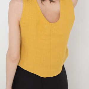 May include: A mustard yellow sleeveless linen top with a scoop neck. The top is cropped and has a slightly curved hemline.