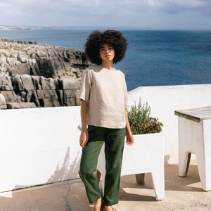 May include: A woman wearing a beige linen top with short sleeves and dark green pants. The outfit is paired with brown sandals. The background features a white wall, ocean, and a cloudy sky. The clothing appears to be casual and comfortable.
