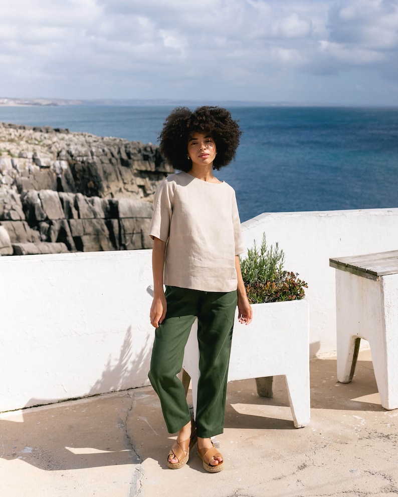 May include: A woman wearing a beige linen top with short sleeves and dark green pants. The outfit is paired with brown sandals. The background features a white wall, ocean, and a cloudy sky. The clothing appears to be casual and comfortable.