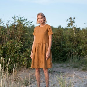 May include: A woman wearing a short-sleeved, rust-colored linen dress with a gathered skirt. She is standing in a grassy area with a wooden walkway behind her.