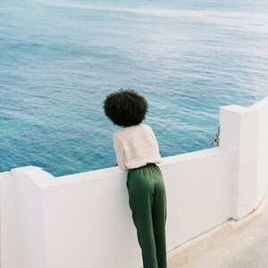May include: A person wearing a cream-colored top and green pants, standing on a white wall, looking out at the ocean. The person is wearing sandals. The ocean is a deep blue color.