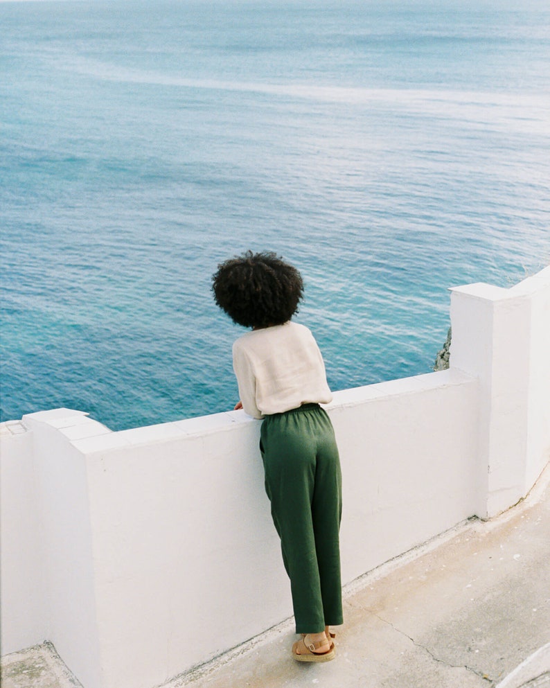 May include: A person wearing a cream-colored top and green pants, standing on a white wall, looking out at the ocean. The person is wearing sandals. The ocean is a deep blue color.