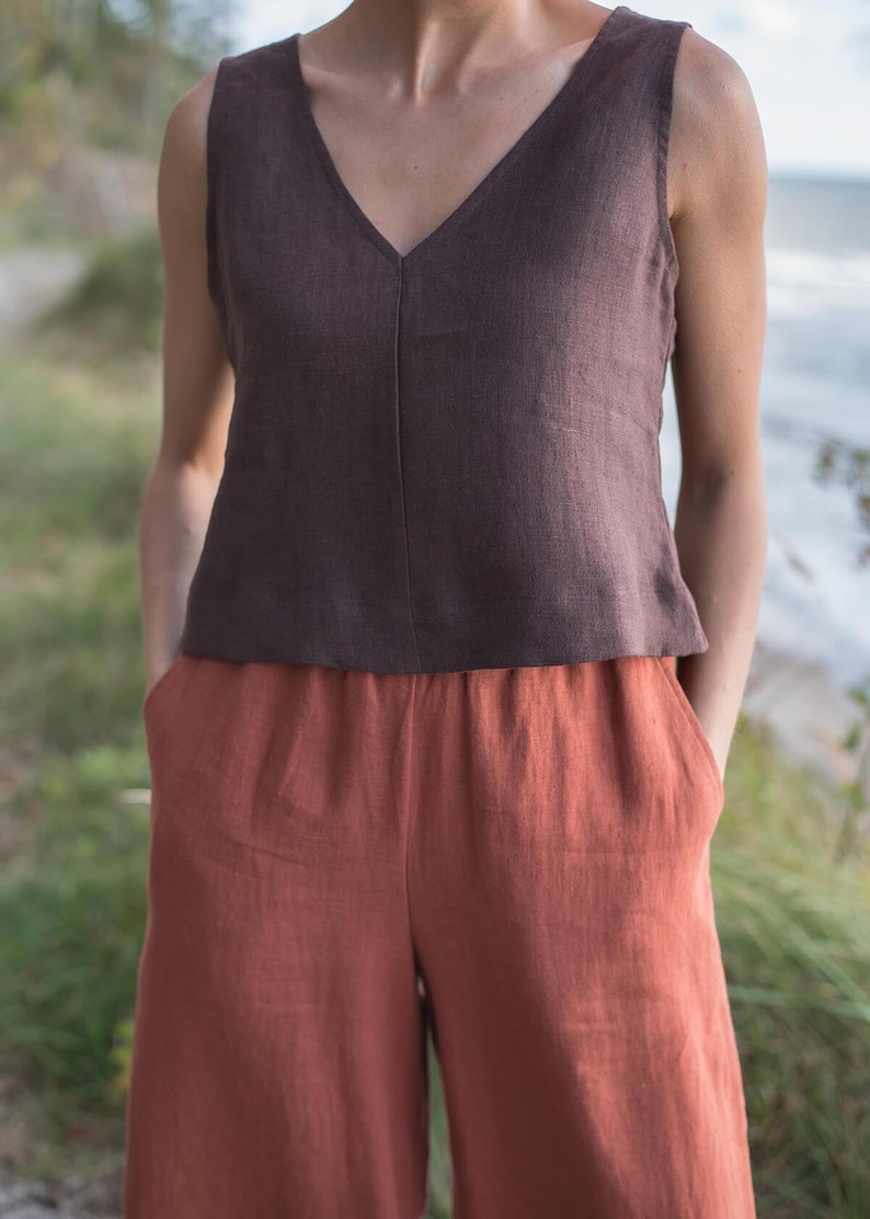 May include: A brown linen sleeveless top with a v-neckline paired with rust-coloured linen trousers. The model is standing outdoors with green grass and a blurred background.