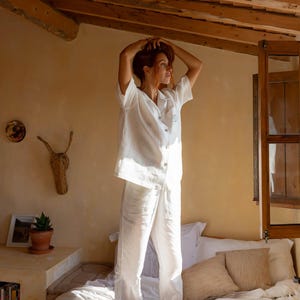 以下が含まれることがあります： ボタン留めのトップスとロングパンツが付いた白いリネンパジャマセット。トップスは襟とシングルボタン留め。パンツはストレートレッグでゆったりとしたフィット感です。