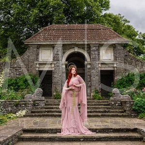 May include: A woman in a pink gown with gold accents stands on a stone staircase in front of a stone archway. The archway is part of a stone building with a tiled roof. The woman is wearing a crown and has red hair. The background is a lush green garden with stone walls.