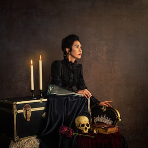 May include: A woman in a black dress sits on a throne-like chair with a skull, a book, and a crown in front of her. Two lit candles are on a black chest behind her.