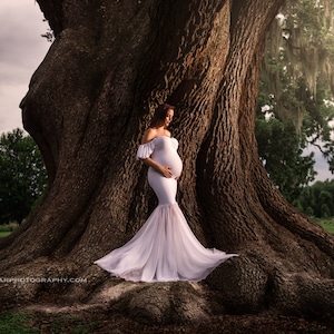 May include: A pregnant woman in a white off-the-shoulder gown stands in front of a large tree with moss hanging from its branches. The woman is holding her belly and looking down.