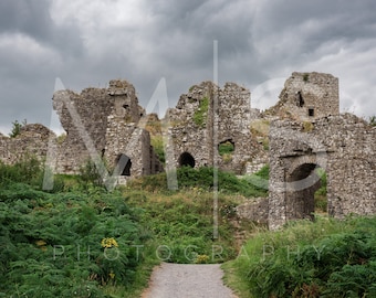 May include: Ruins of a stone castle with a path leading to the entrance. The castle is overgrown with vegetation and the sky is cloudy.