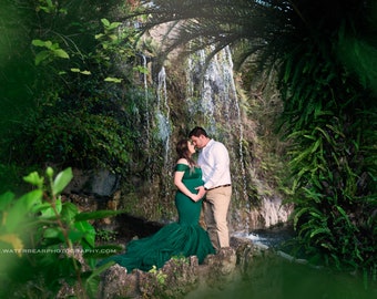 May include: A pregnant woman in a long, emerald green gown stands with her partner in front of a waterfall. They are both looking at each other and smiling. The waterfall is surrounded by lush greenery.