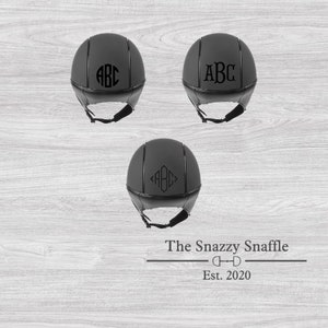 May include: Three black riding helmets with a black monogram of the letters ABC on each. The helmets are on a woodgrain background. The text "The Snazzy Snaffle" and "Est. 2020" is below the helmets.