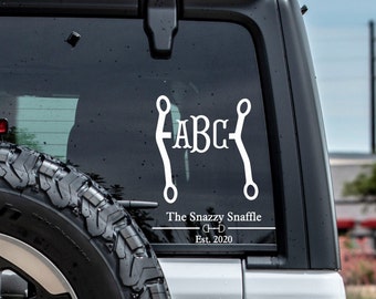 May include: White vinyl decal with a snaffle bit design and the letters "ABC" inside the bit. The text "The Snazzy Snaffle" and "Est. 2020" is below the bit.