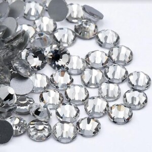 May include: A collection of clear, faceted rhinestones with a flat back. The rhinestones are oval and round, reflecting light. The image shows a variety of sizes and shapes, suitable for crafts and embellishments.