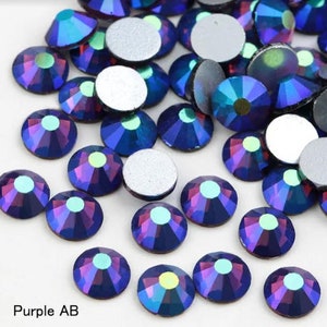 May include: A collection of iridescent purple AB rhinestones. Each rhinestone is round with a faceted surface, reflecting shades of blue, purple, and green. The text "Purple AB" is visible at the bottom left.