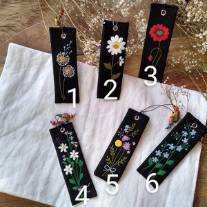 K&ouml;nnte beinhalten: Sechs schwarze Stoff-Lesezeichen mit gestickten Blumenmotiven. Jedes Lesezeichen hat eine wei&szlig;e Nummer von 1 bis 6 in der unteren rechten Ecke.