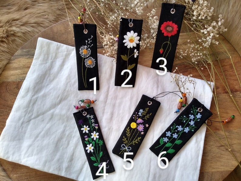 K&ouml;nnte beinhalten: Sechs schwarze Stoff-Lesezeichen mit gestickten Blumenmotiven. Jedes Lesezeichen hat eine wei&szlig;e Nummer von 1 bis 6 in der unteren rechten Ecke.