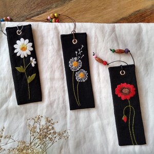 K&ouml;nnte beinhalten: Drei schwarze Stoff-Lesezeichen mit gestickten Blumen. Das erste Lesezeichen hat eine wei&szlig;e G&auml;nsebl&uuml;mchen, das zweite hat einen wei&szlig;en L&ouml;wenzahn und das dritte hat eine rote Mohnblume.