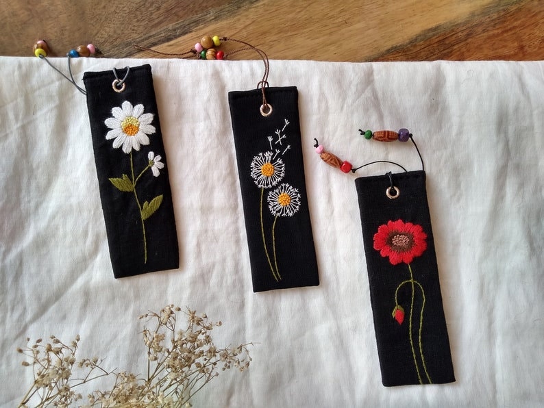 K&ouml;nnte beinhalten: Drei schwarze Stoff-Lesezeichen mit gestickten Blumen. Das erste Lesezeichen hat eine wei&szlig;e G&auml;nsebl&uuml;mchen, das zweite hat einen wei&szlig;en L&ouml;wenzahn und das dritte hat eine rote Mohnblume.