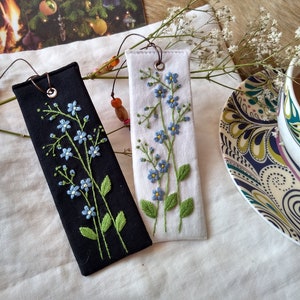K&ouml;nnte beinhalten: Zwei handgefertigte Stoff-Lesezeichen mit gestickten Vergissmeinnicht-Blumen. Ein Lesezeichen ist schwarz mit wei&szlig;en Blumen und das andere ist wei&szlig; mit blauen Blumen.
