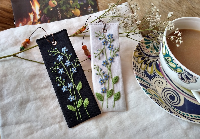 K&ouml;nnte beinhalten: Zwei handgefertigte Stoff-Lesezeichen mit gestickten Vergissmeinnicht-Blumen. Ein Lesezeichen ist schwarz mit wei&szlig;en Blumen und das andere ist wei&szlig; mit blauen Blumen.