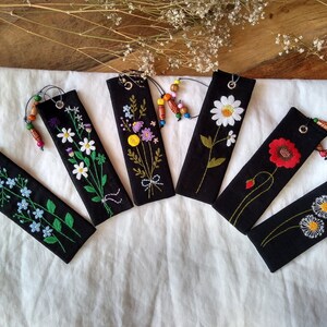 K&ouml;nnte beinhalten: Sechs schwarze Stoff-Lesezeichen mit gestickten Blumenmotiven. Jedes Lesezeichen zeigt eine andere Blume, darunter G&auml;nsebl&uuml;mchen, Vergissmeinnicht und eine Mohnblume. Die Lesezeichen haben einen kleinen Metallring und einen Perlenanh&auml;nger.