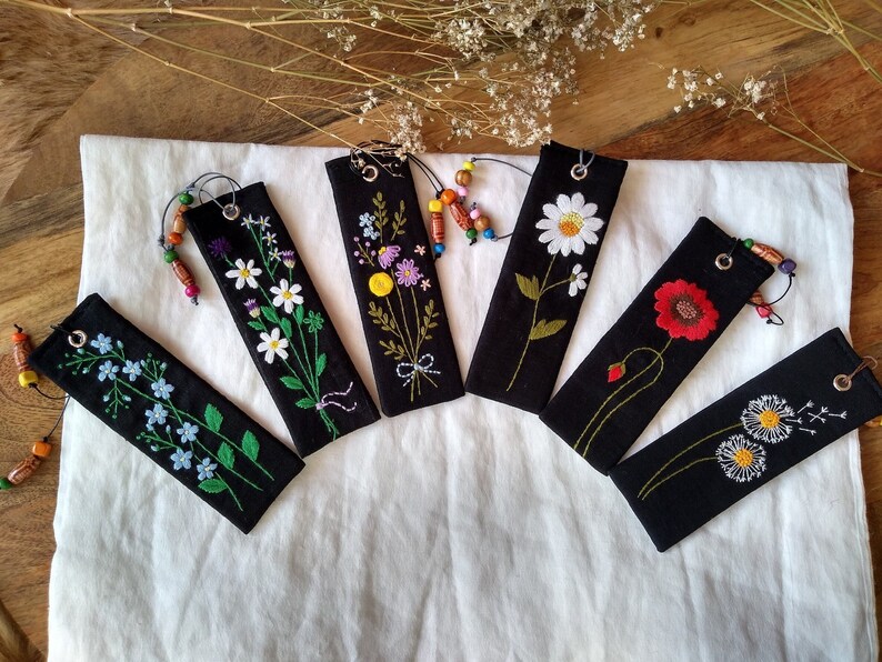 K&ouml;nnte beinhalten: Sechs schwarze Stoff-Lesezeichen mit gestickten Blumenmotiven. Jedes Lesezeichen zeigt eine andere Blume, darunter G&auml;nsebl&uuml;mchen, Vergissmeinnicht und eine Mohnblume. Die Lesezeichen haben einen kleinen Metallring und einen Perlenanh&auml;nger.