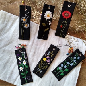 K&ouml;nnte beinhalten: Sechs schwarze Stoff-Lesezeichen mit bunten gestickten Blumen. Die Lesezeichen haben eine schwarze Schnur, die oben mit Holzperlen befestigt ist.