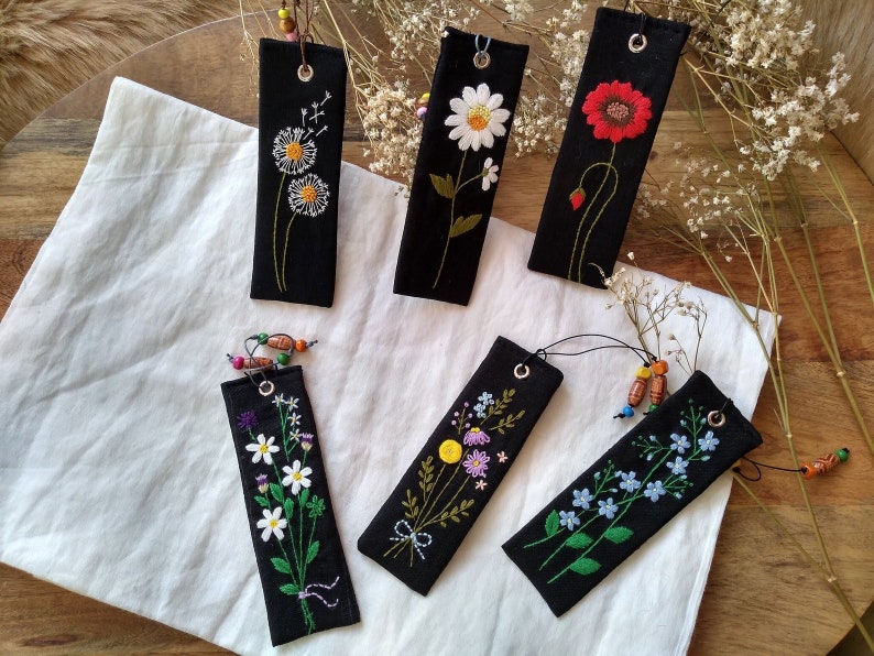 K&ouml;nnte beinhalten: Sechs schwarze Stoff-Lesezeichen mit bunten gestickten Blumen. Die Lesezeichen haben eine schwarze Schnur, die oben mit Holzperlen befestigt ist.