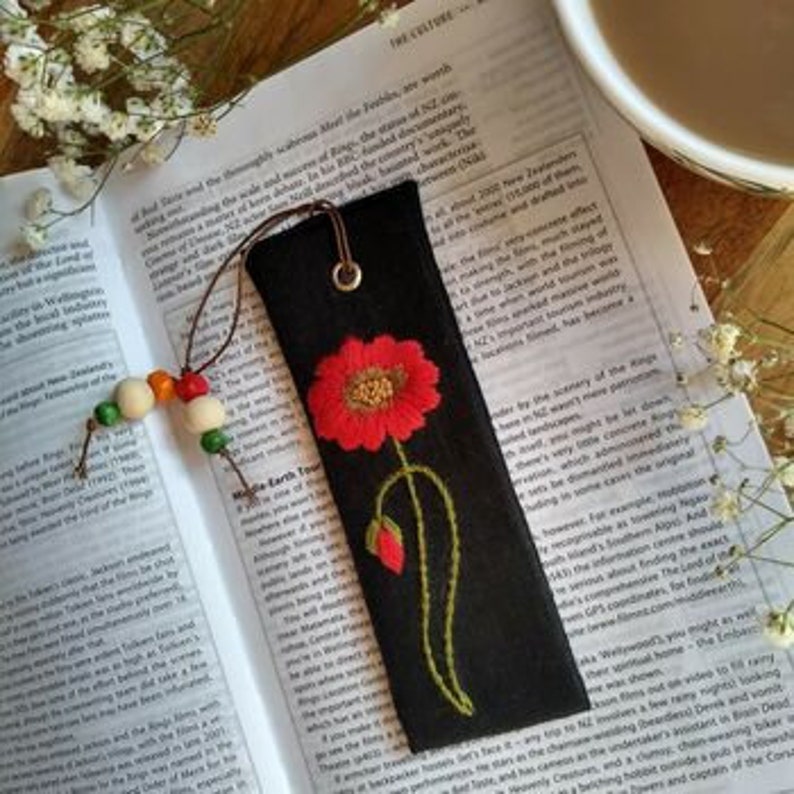 K&ouml;nnte beinhalten: Ein schwarzes Stoff Lesezeichen mit einer aufgestickten roten Mohnblume. Das Lesezeichen hat einen braunen Lederanh&auml;nger und eine Schnur aus Holzperlen.