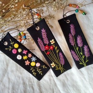 K&ouml;nnte beinhalten: Drei schwarze Stoff-Lesezeichen mit gestickten Blumenmotiven. Ein Lesezeichen zeigt ein Lavendel-Design, ein anderes eine Vielzahl von bunten Blumen und das dritte ein schlichtes Blumenmuster mit gelben und gr&uuml;nen Akzenten.