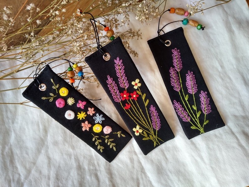 K&ouml;nnte beinhalten: Drei schwarze Stoff-Lesezeichen mit gestickten Blumenmotiven. Ein Lesezeichen zeigt ein Lavendel-Design, ein anderes eine Vielzahl von bunten Blumen und das dritte ein schlichtes Blumenmuster mit gelben und gr&uuml;nen Akzenten.