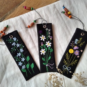 K&ouml;nnte beinhalten: Drei schwarze Stoff-Lesezeichen mit gestickten Blumenmustern. Jedes Lesezeichen hat ein anderes Blumenmuster und ist mit einem bunten Perlen-Quast verziert.