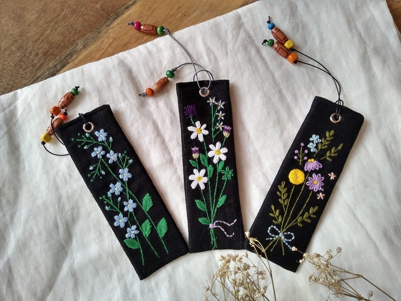 K&ouml;nnte beinhalten: Drei schwarze Stoff-Lesezeichen mit gestickten Blumenmustern. Jedes Lesezeichen hat ein anderes Blumenmuster und ist mit einem bunten Perlen-Quast verziert.