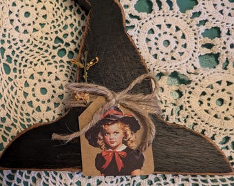 May include: A rustic, black wooden witch hat decoration. It features a vintage image of a girl in a black dress and hat, tied with twine and a small sprig of dried flowers. The hat is set against a white lace background.