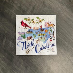 May include: White paper napkin with a watercolor illustration of North Carolina. The design includes a red cardinal, a bear, a pig, a horse, and the text "North Carolina" in blue script. The napkin is square and has a textured surface.