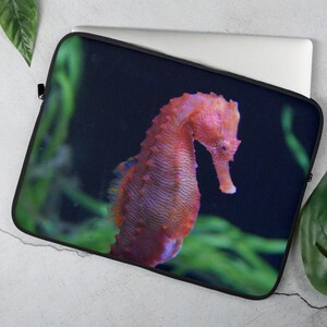 Peut inclure: Une housse d'ordinateur portable avec un motif d'hippocampe orange vif sur fond bleu foncé et des plantes aquatiques vertes. L'hippocampe présente des textures détaillées et est au centre du design. La housse est noire.