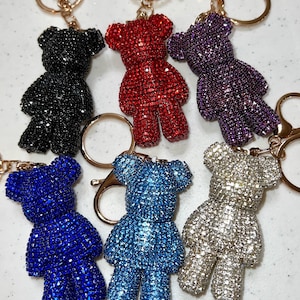 Pode incluir: Seis chaveiros de urso de pelúcia cobertos de strass em várias cores: preto, vermelho, roxo, azul, azul claro e prata. Cada urso tem um chaveiro e um fecho dourados.