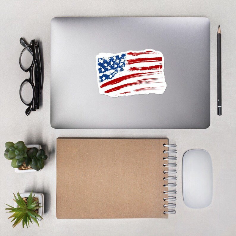 May include: A distressed American flag sticker on a silver laptop computer. The sticker is white with red and blue paint splatters.