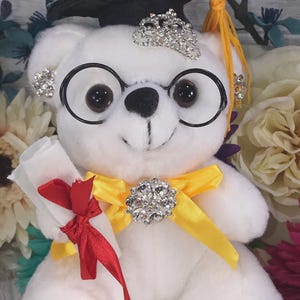 May include: A white plush teddy bear wearing a black graduation cap, a rhinestone tiara, and round glasses. The bear is holding a rolled-up diploma with a red ribbon and is wearing a yellow ribbon with a rhinestone brooch.